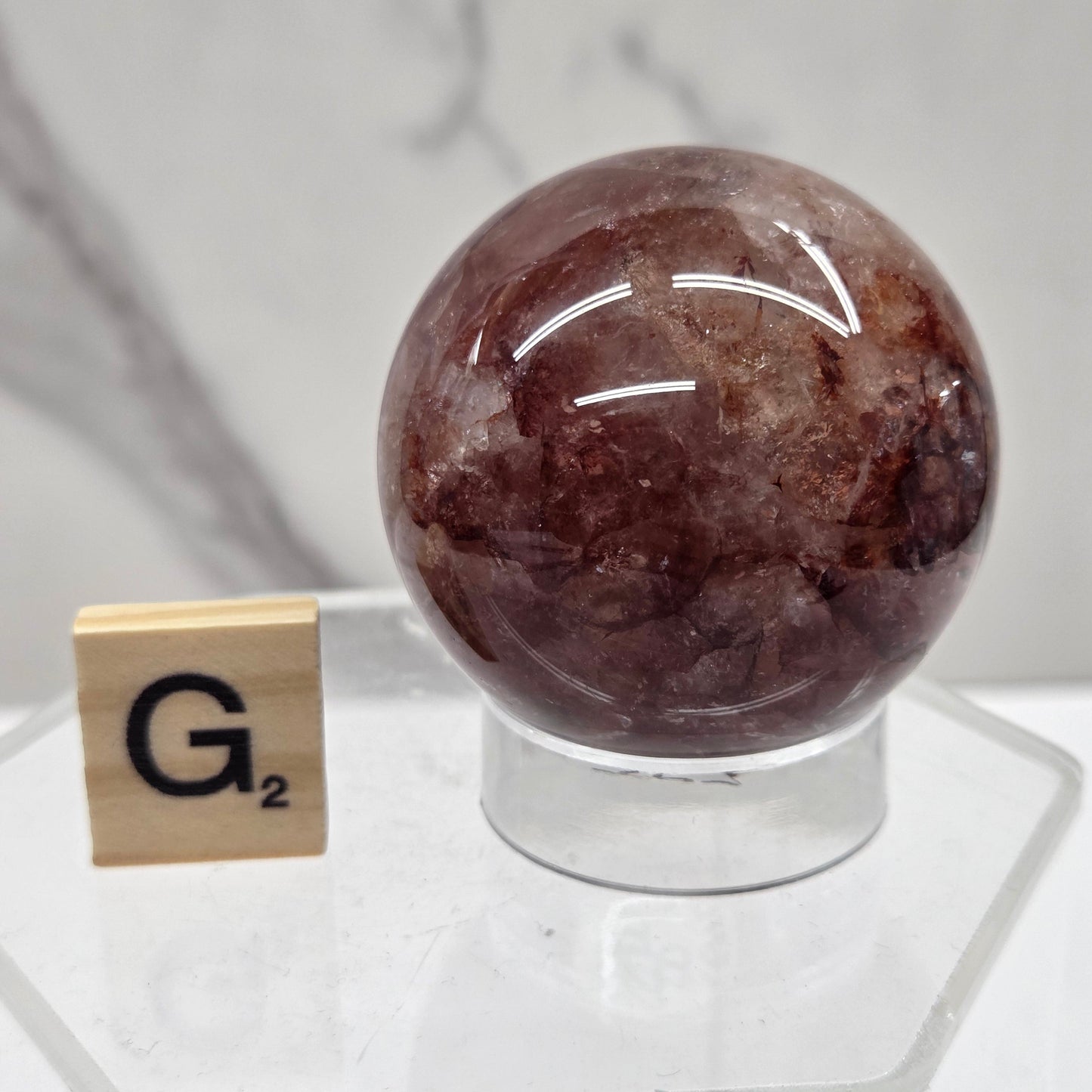 A polished brown gemstone sphere with natural crystalline inclusions, mounted on a clear acrylic stand with a small wooden tile displaying 'G²' in front, set against a light marble-patterned background.
