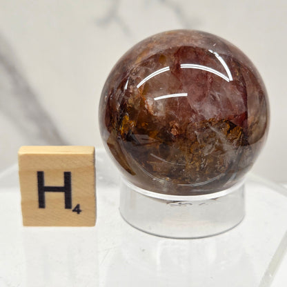 A polished brown and amber crystal sphere with inclusions, mounted on a clear acrylic stand next to a small wooden tile with a black letter H, on a light marble surface.