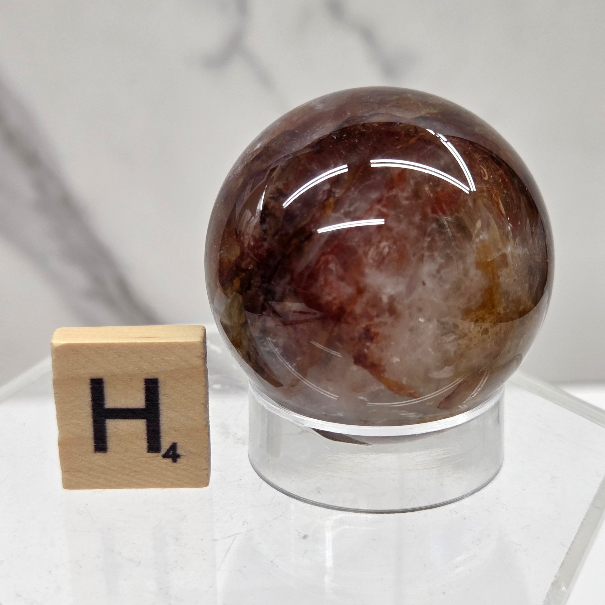 A polished brown and smoky translucent stone sphere mounted on a clear acrylic stand, with a small wooden tile marked H4 placed below it on a light marble-like surface.