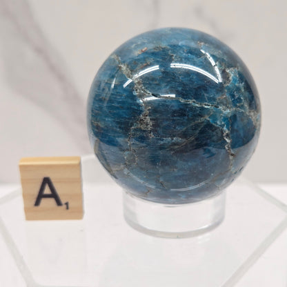 A polished blue stone sphere with natural white veining, mounted on a clear acrylic stand with a small wooden letter tile marked 'A' below it, set against a light marble-patterned background.