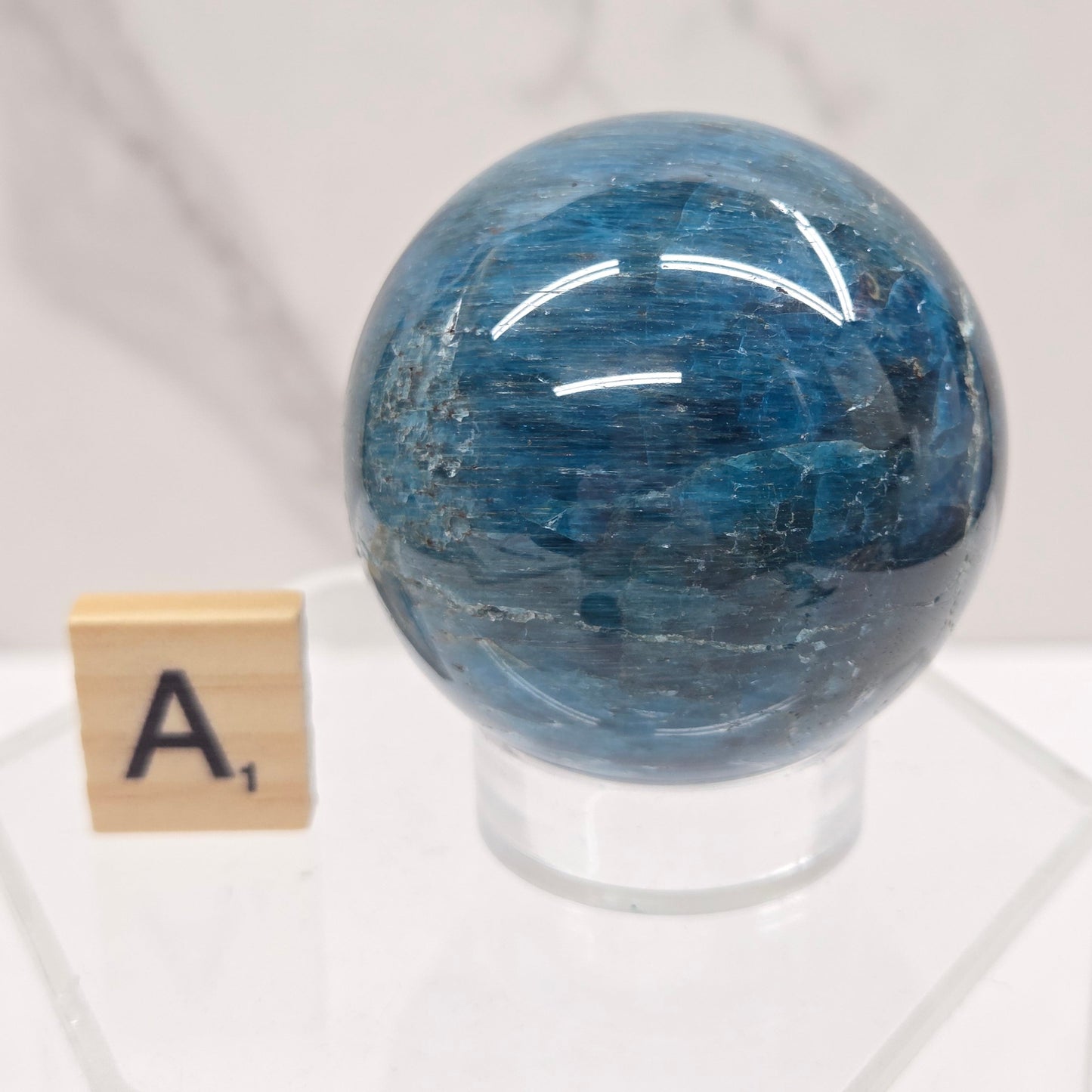 A polished blue stone sphere mounted on a clear acrylic stand, with a small wooden letter tile marked 'A' below it, set against a light marble-patterned background.