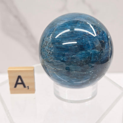 A polished blue stone sphere mounted on a clear acrylic stand, with a small wooden letter tile marked 'A' below it, set against a light marble-patterned background.