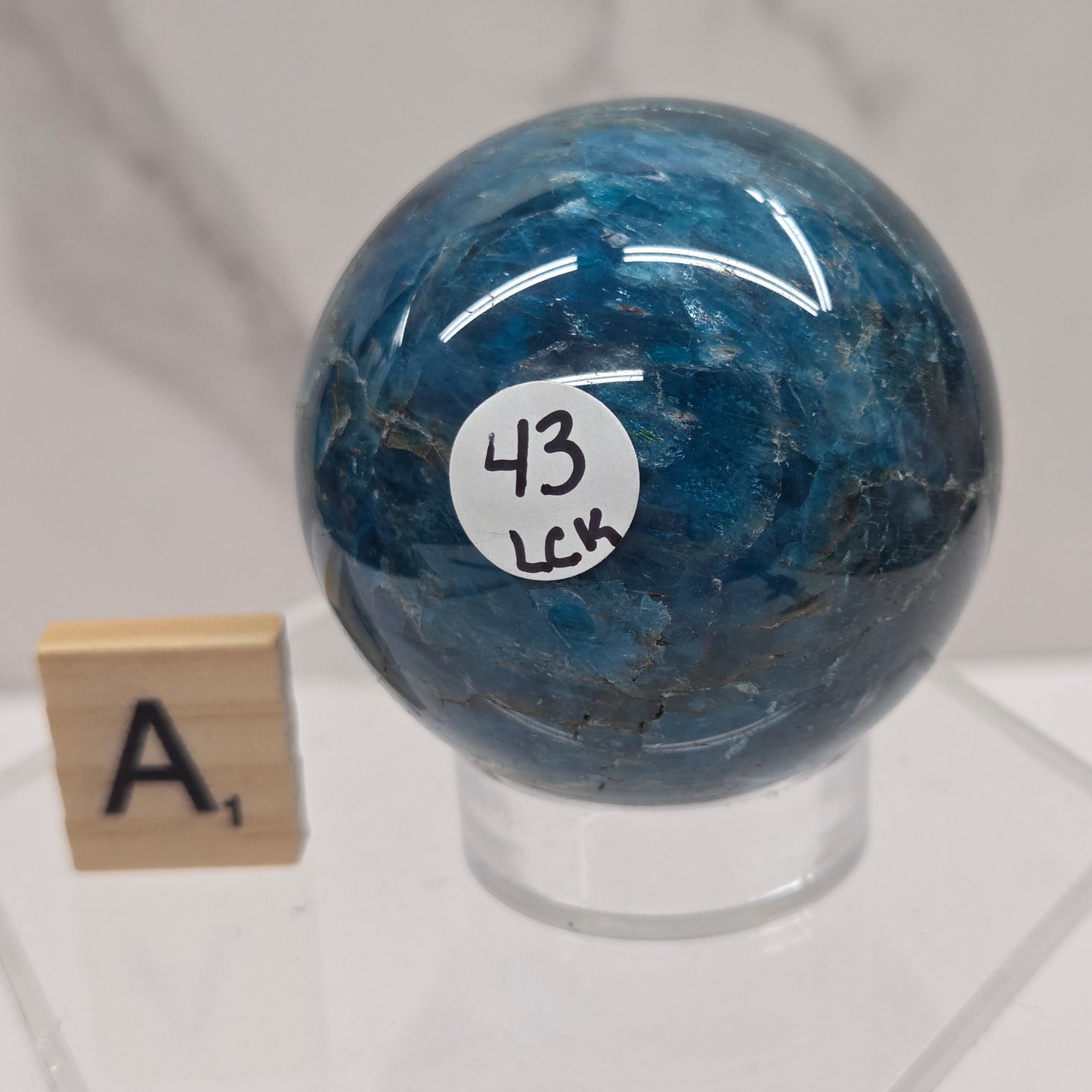 A polished blue stone sphere with natural marbling, mounted on a clear acrylic stand. A small white sticker with the number 43 is attached to the sphere, and a wooden letter tile stands below on the stand.