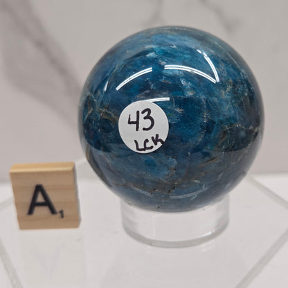 A polished blue stone sphere with natural marbling, mounted on a clear acrylic stand. A small white sticker with the number 43 is attached to the sphere, and a wooden letter tile stands below on the stand.