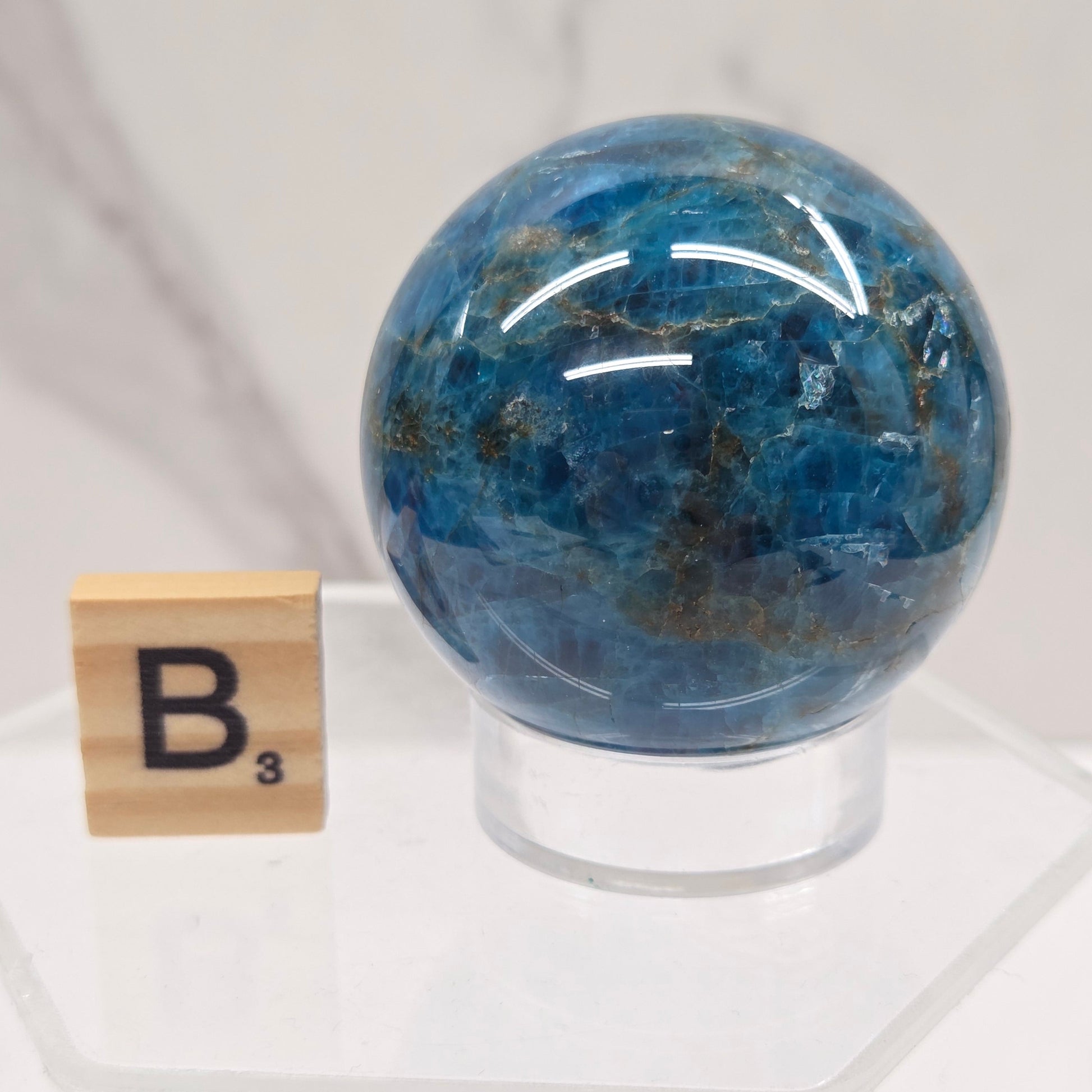 A polished blue stone sphere mounted on a clear acrylic stand, with a small wooden tile marked 'B' placed below it, set against a light marble-like background.
