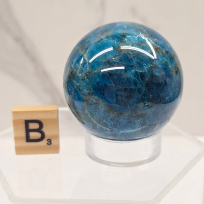 A polished blue gemstone sphere with natural marbling, displayed on a clear acrylic stand next to a small wooden tile labeled B3, against a light marble background.