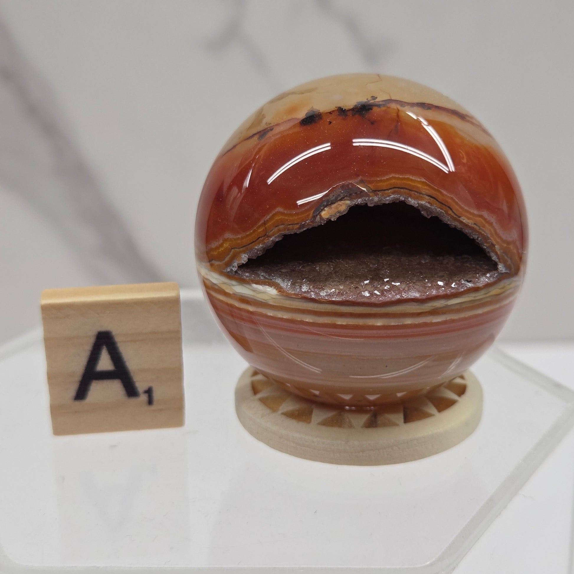 A carnelian sphere, made from brown and orange banded agate with a dark interior, displayed on a small wooden base next to a Scrabble tile showing 'A1'.