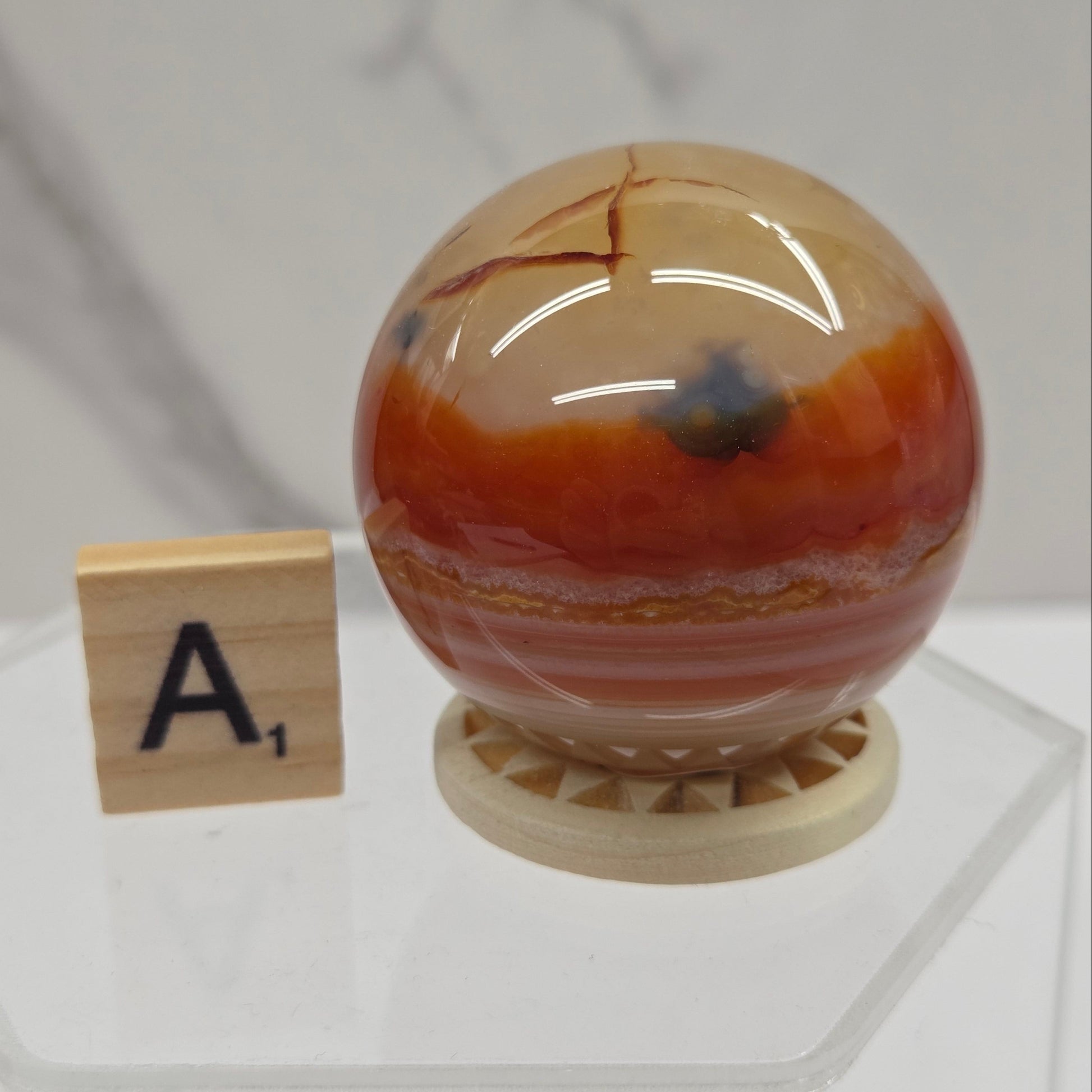 A polished round agate crystal sphere with orange, brown, and beige banded patterns sits on a small wooden base next to a wooden letter tile displaying 'A1'.