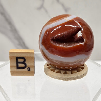 A round carnelian sphere, displayed next to a wooden letter block on a white marble surface.