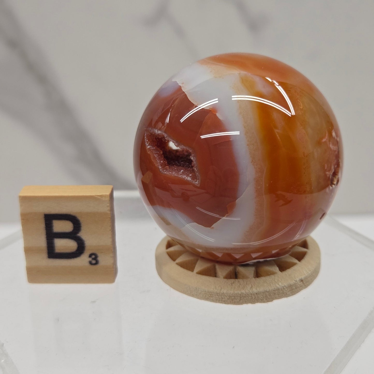 A polished agate crystal sphere with swirling bands of orange, brown, white, and reddish tones, displayed on a small wooden stand next to a wooden letter tile on a clear acrylic platform.
