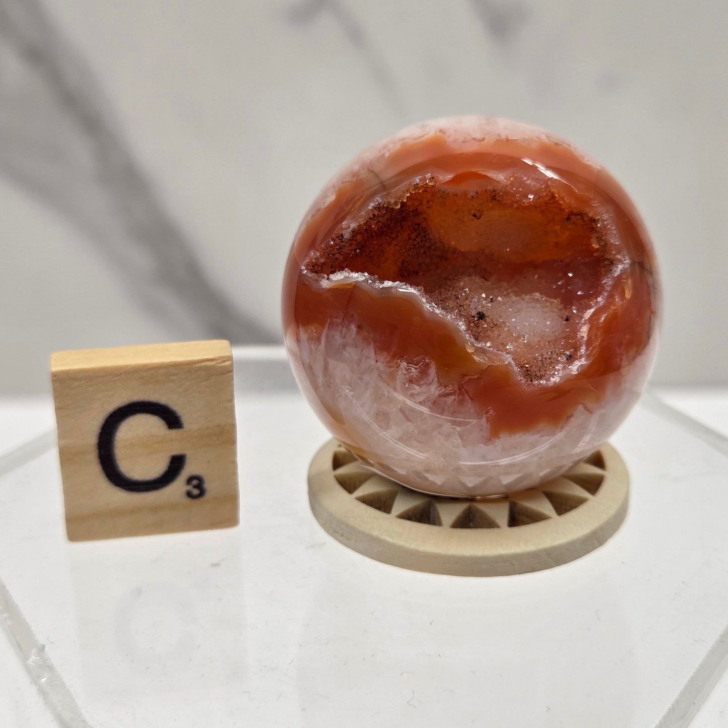 A polished orange-brown agate geode sphere on a small wooden display base, shown next to a wooden Scrabble tile marked C3 on a clear acrylic stand against a light marble-patterned background.