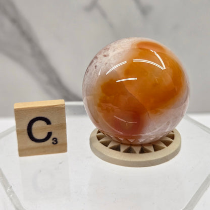 A polished orange and white agate crystal ball resting on a small round wooden stand, displayed next to a wooden Scrabble tile marked C3 on a clear acrylic surface against a light marble-look background.
