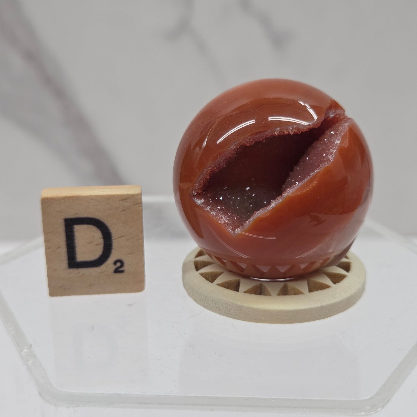 A glossy reddish-brown geode-like decorative sphere with a hollow center, displayed on a small round stand next to a wooden Scrabble tile showing the letter D2, set on a clear acrylic surface against a light marble-pattern background.