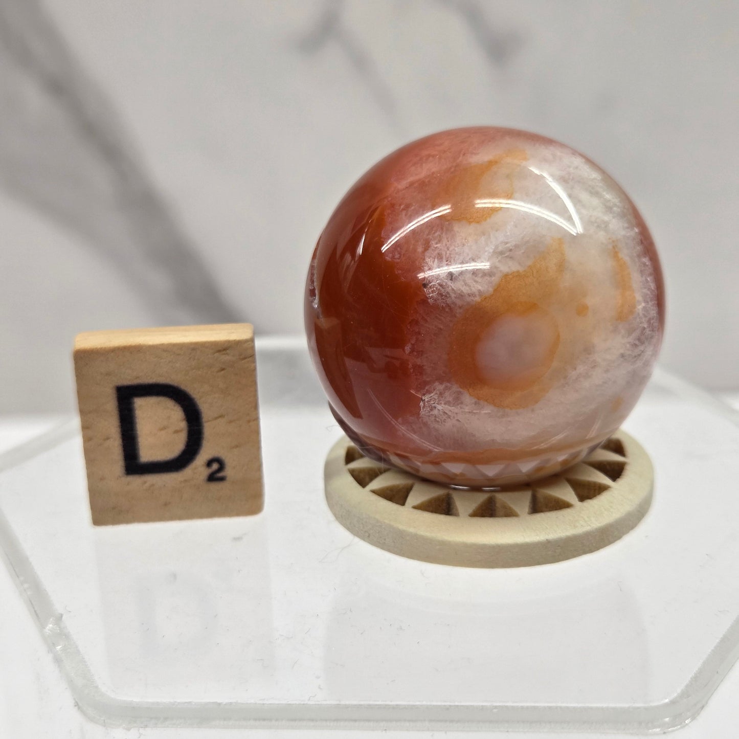 A polished orange and white agate crystal ball resting on a small wooden display stand, shown next to a Scrabble tile on a clear acrylic surface against a light marble-patterned background.