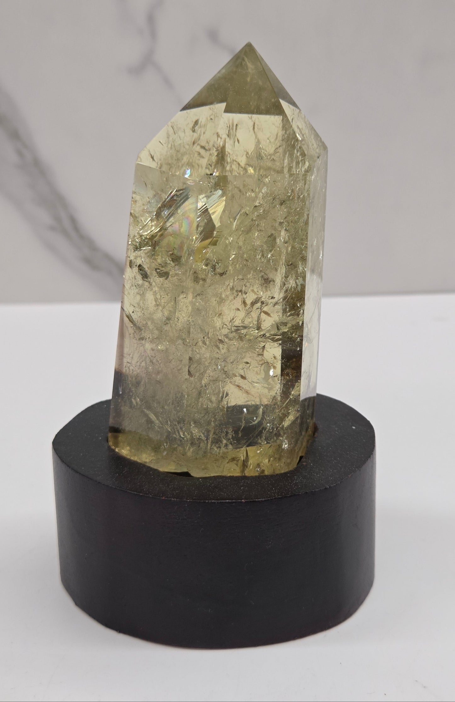 A natural clear quartz crystal point with internal inclusions, mounted on a cylindrical black display stand against a white and gray marble-patterned background.