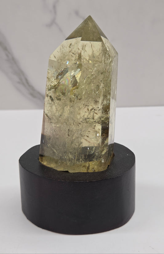 A natural clear quartz crystal point with internal inclusions, mounted on a cylindrical black display stand against a white and gray marble-patterned background.