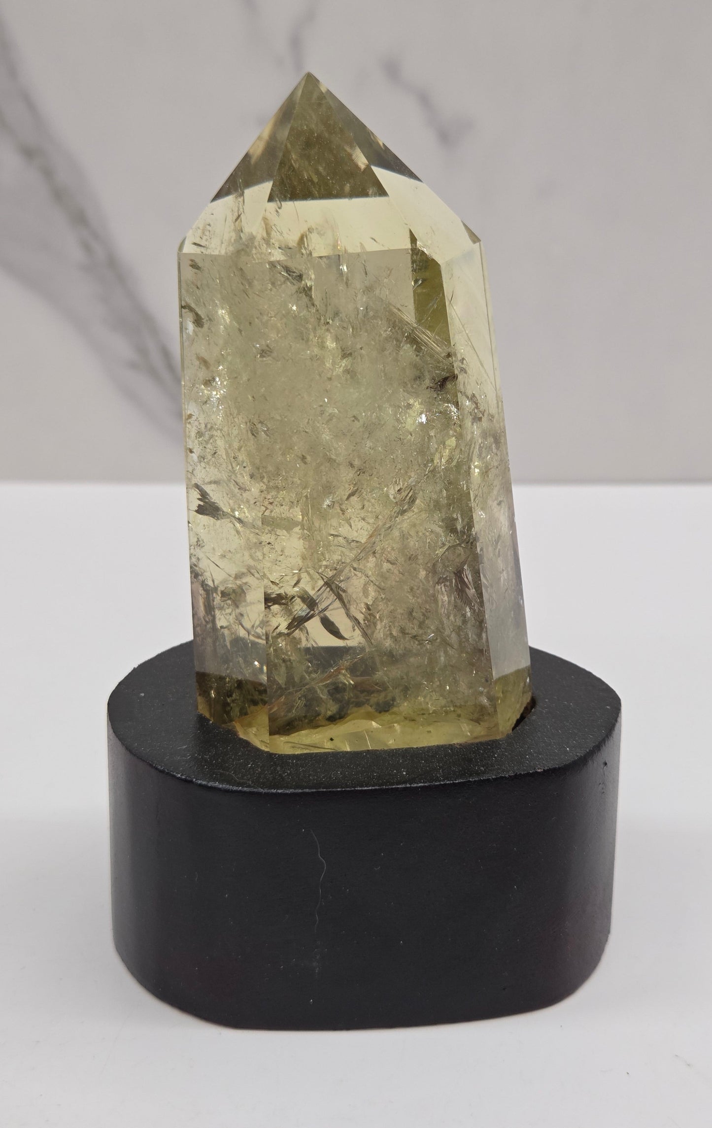A faceted natural crystal point with a pale yellow-green hue mounted on a cylindrical black display stand against a white background.