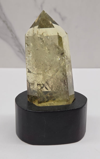 A faceted natural crystal point with a pale yellow-green hue mounted on a cylindrical black display stand against a white background.