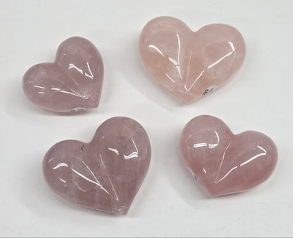 Four polished heart-shaped rose quartz gemstones in varying shades of dusty pink and lavender, arranged on a plain white background.
