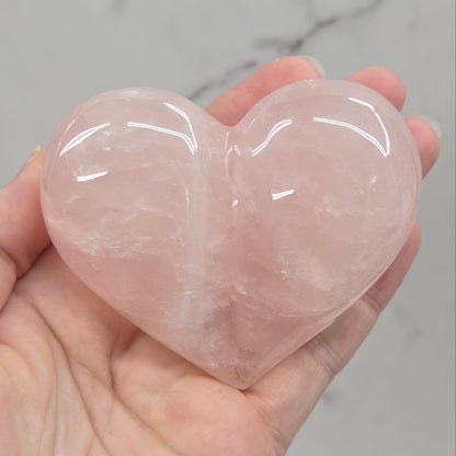 A hand holding a polished rose quartz heart-shaped crystal against a light gray marble background.