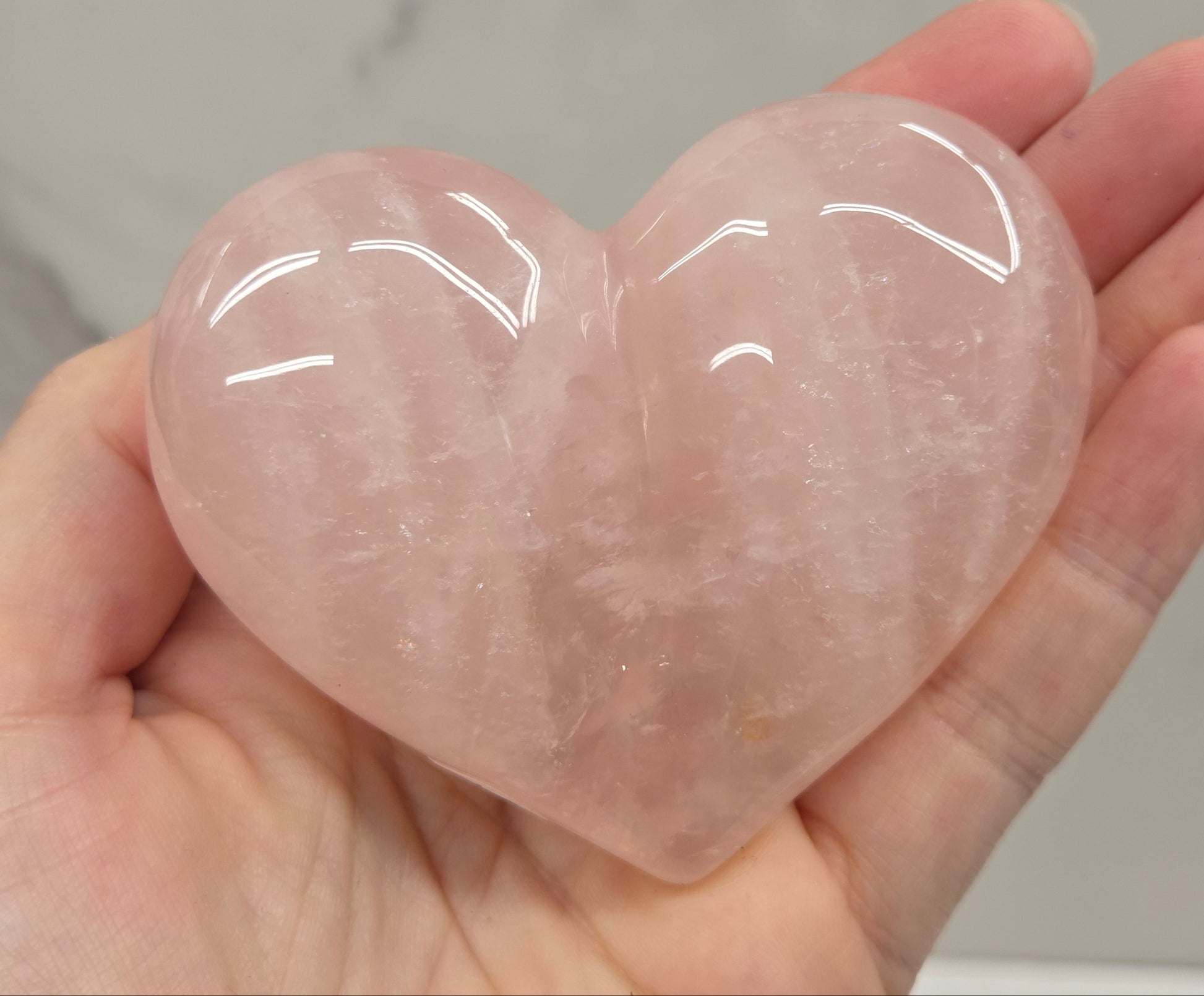 A polished rose quartz heart-shaped crystal held in a person's hand against a light marble background.