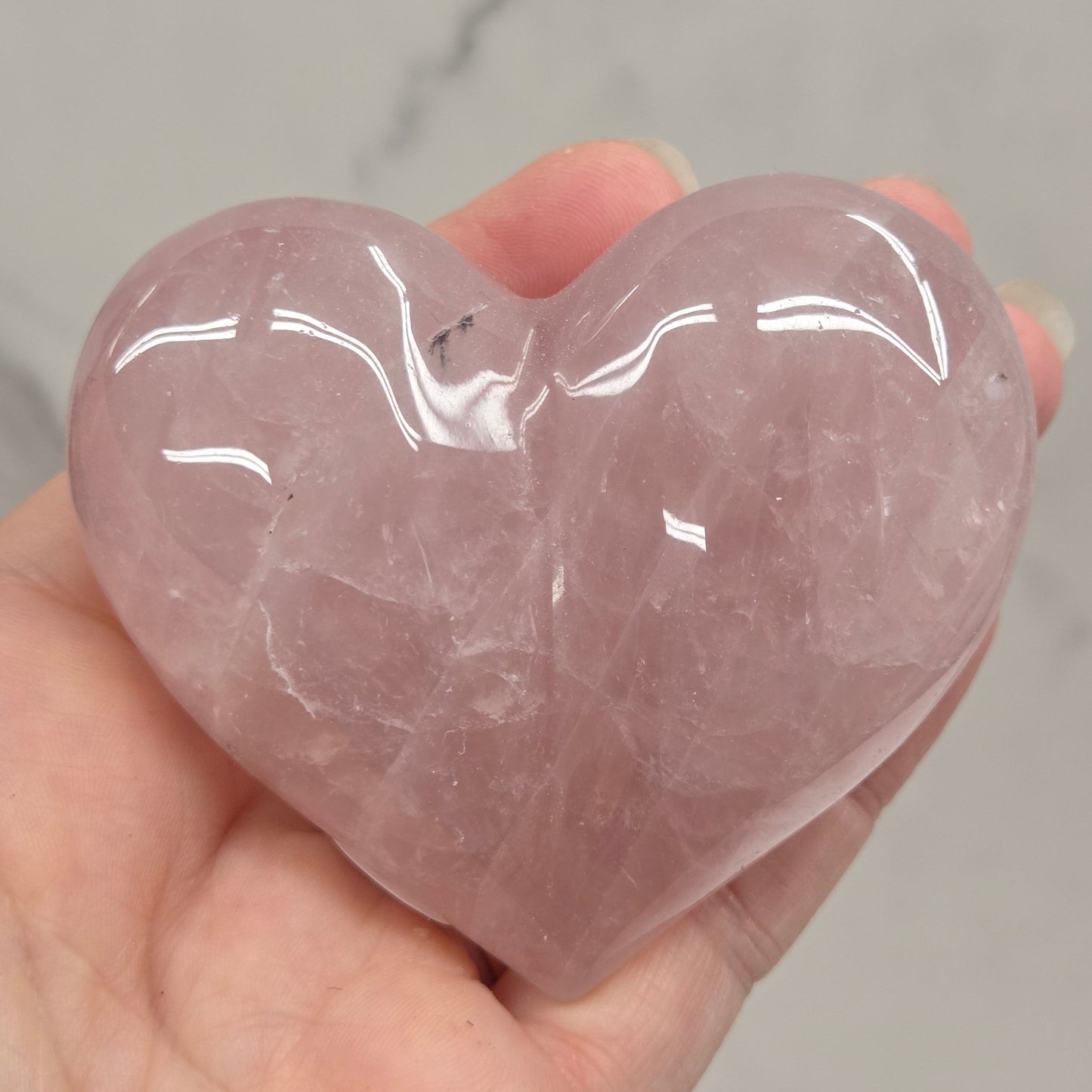 A hand holding a polished rose quartz heart-shaped crystal against a light gray marble background.