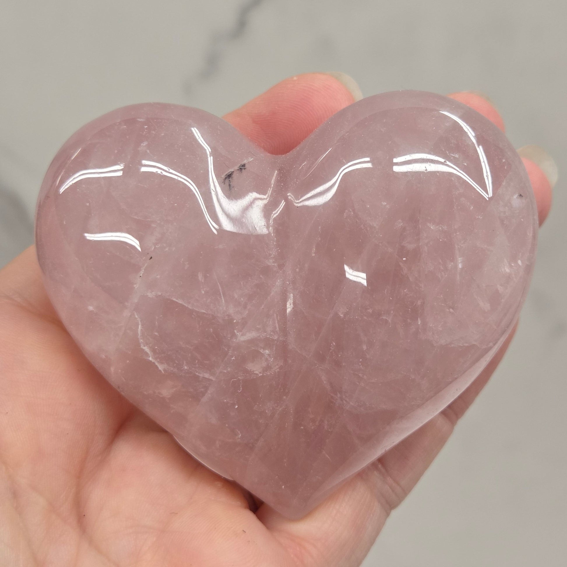 A hand holding a polished rose quartz heart-shaped crystal against a light gray marble background.