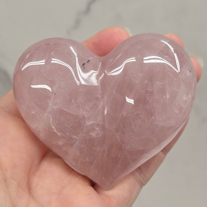 A hand holding a polished rose quartz heart-shaped crystal against a light gray marble background.