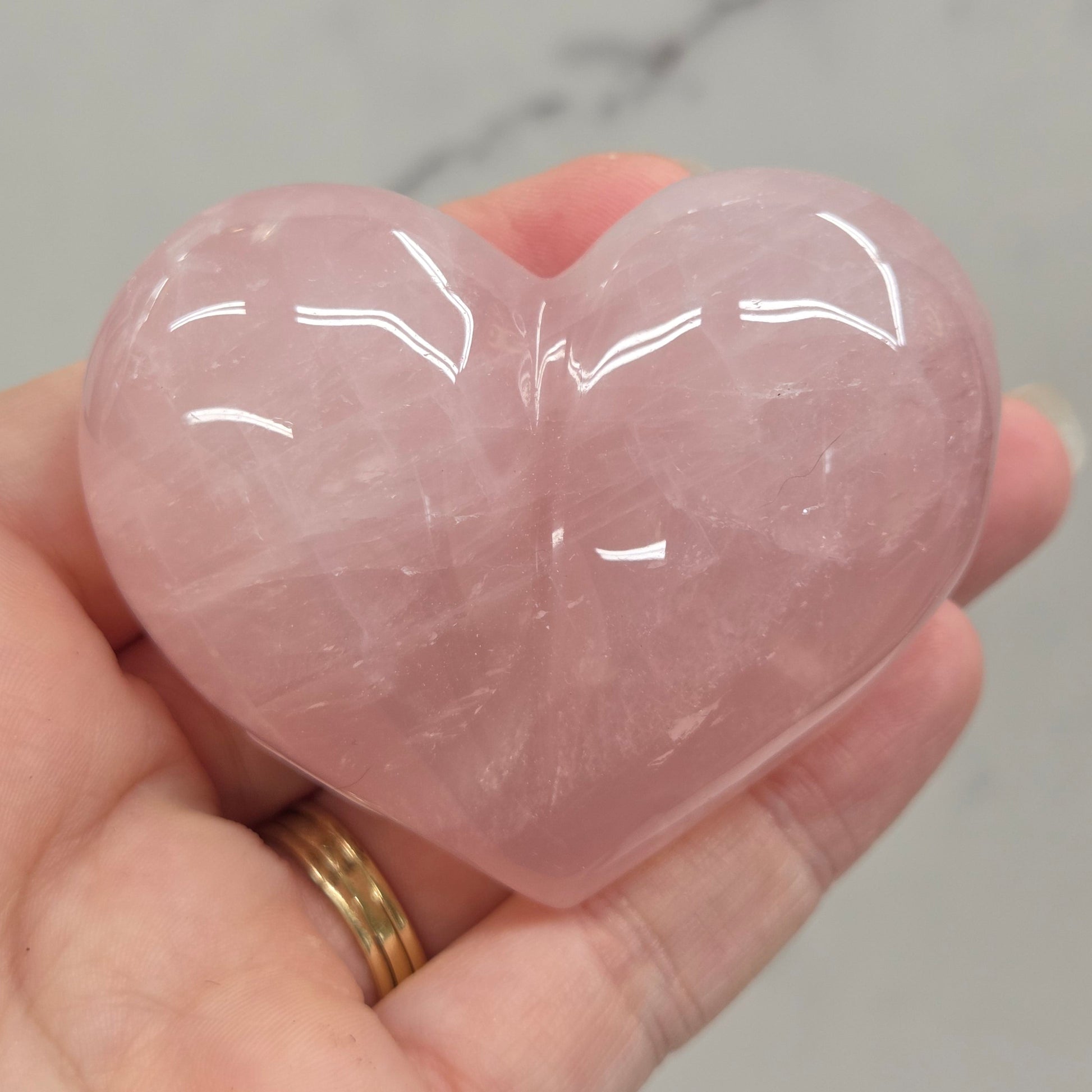 A hand holding a polished pink rose quartz heart-shaped crystal against a light gray marble background.