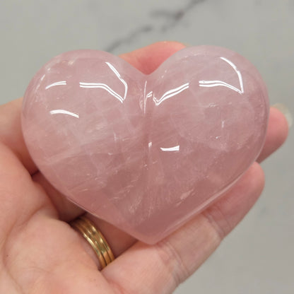 A hand holding a polished pink rose quartz heart-shaped crystal against a light gray marble background.