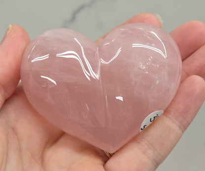 A hand holding a polished, heart-shaped rose quartz crystal against a light gray marble background.