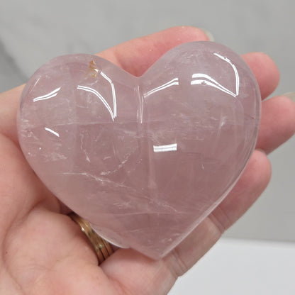 A hand holding a polished rose quartz heart-shaped crystal against a light gray marble background.