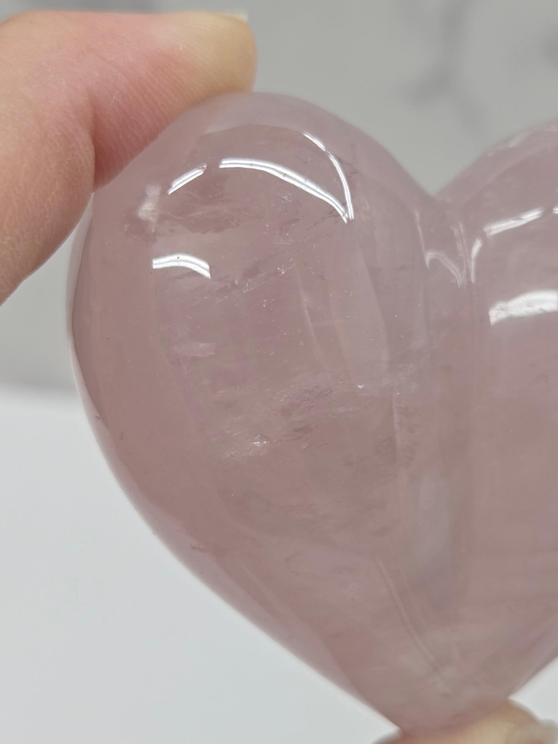 A close-up of a hand holding a polished rose quartz crystal heart against a light gray background.