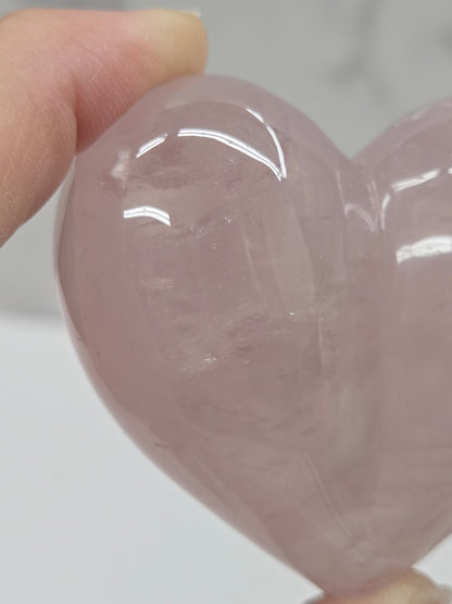 A close-up of a hand holding a polished rose quartz crystal heart against a light gray background.