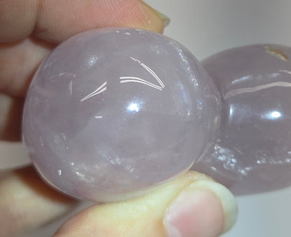 A close-up of a person holding a double-gourd-shaped carved stone pendant made of pale lavender quartzite, showing its smooth, polished texture and natural inclusions.