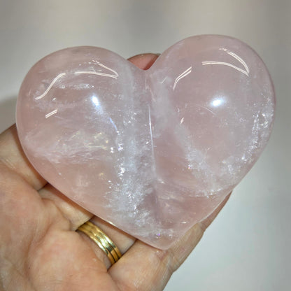 A heart-shaped rose quartz crystal held in a person's hand, showing its soft pink color and natural cloudy texture.