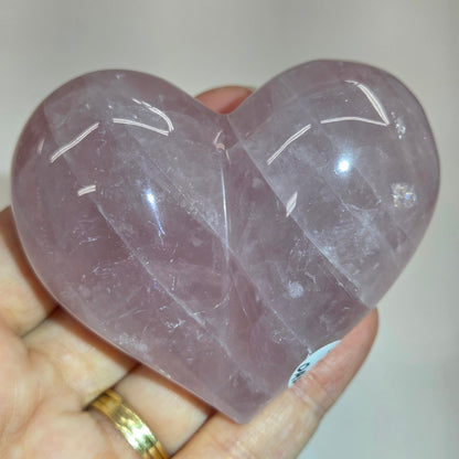 A heart-shaped rose quartz crystal ball held in a person's hand, showing its smooth, polished surface and natural lavender-purple color.