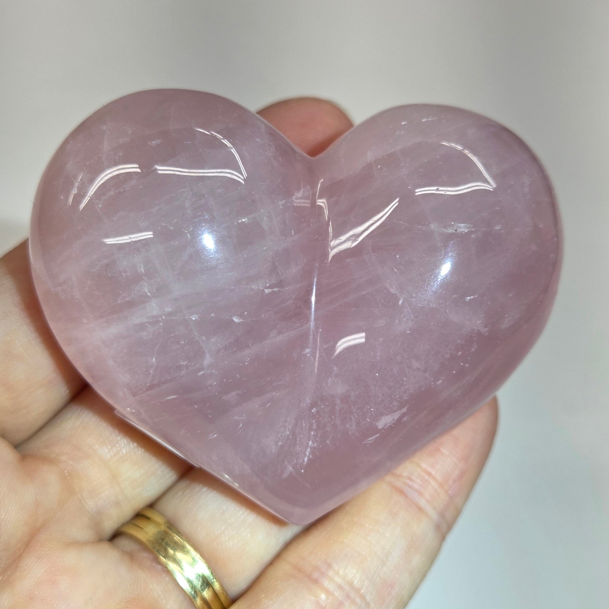 A heart-shaped polished pink stone held in a person's hand, with a gold ring visible on one finger against a plain light background.