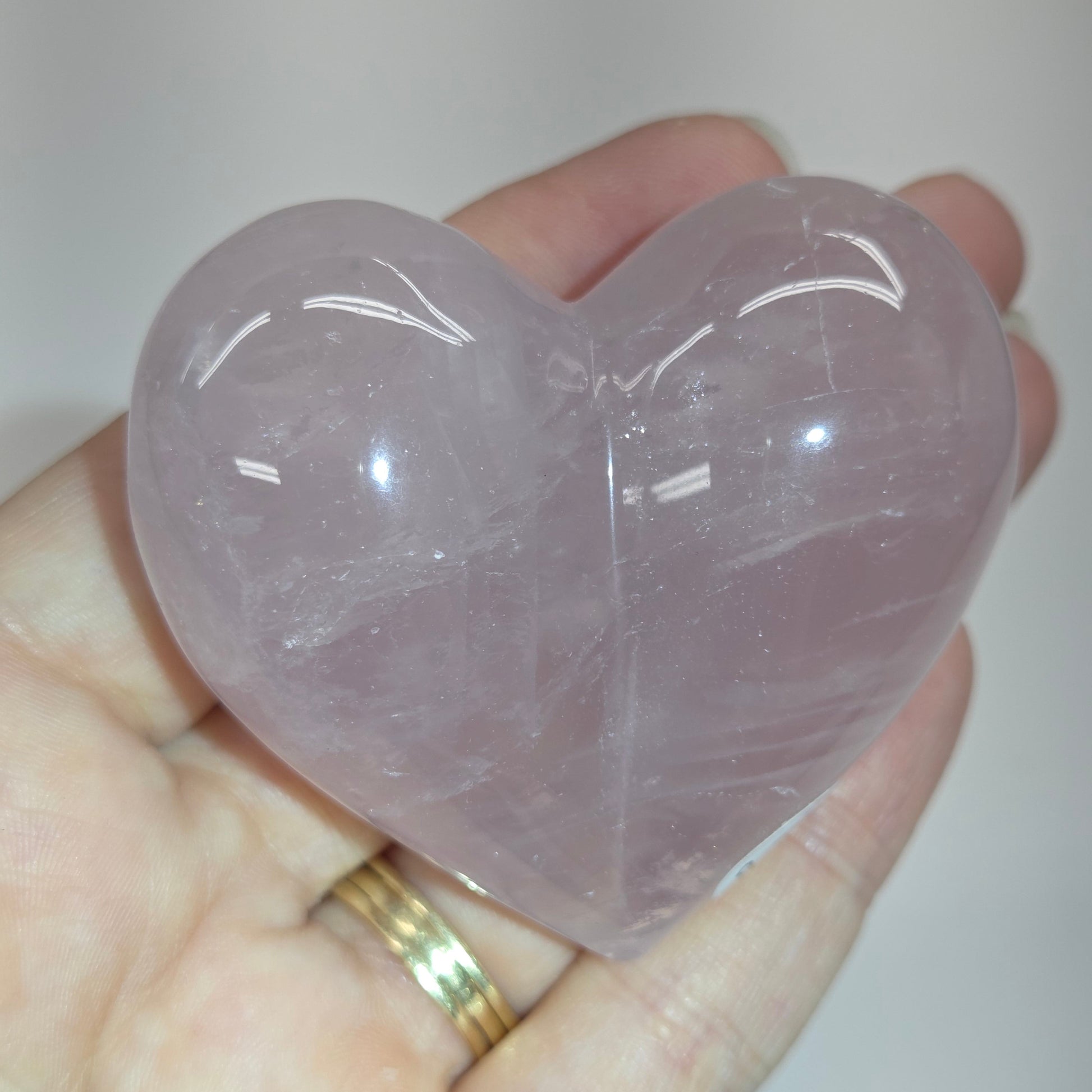 A heart-shaped rose quartz crystal ball held in a person's hand. The polished stone has a soft lavender-pink color with natural internal inclusions and smooth, reflective surfaces.