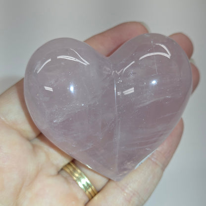 A heart-shaped rose quartz crystal ball held in a person's hand. The polished stone has a soft lavender-pink color with natural internal inclusions and smooth, reflective surfaces.