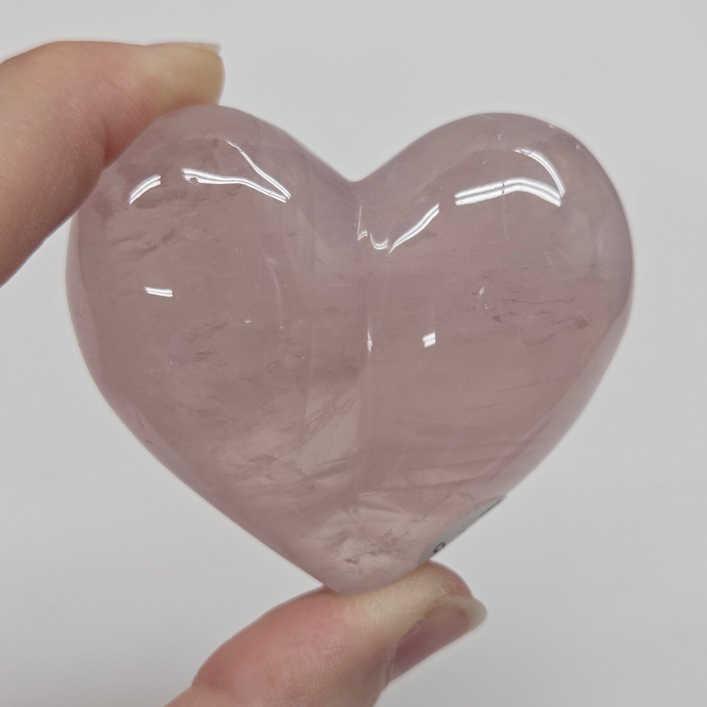 A hand holding a polished, heart-shaped rose quartz crystal against a plain light gray background.