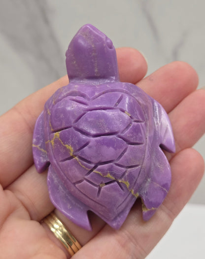 A carved purple turtle figurine with gold veining, held in a person's hand against a light gray marble background.