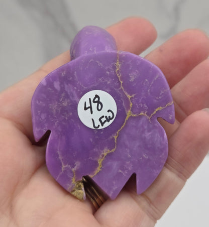 A hand holding a purple polished stone object with natural gold veining and a small white sticker labeled '43 $20' on it, against a light marble-like background.