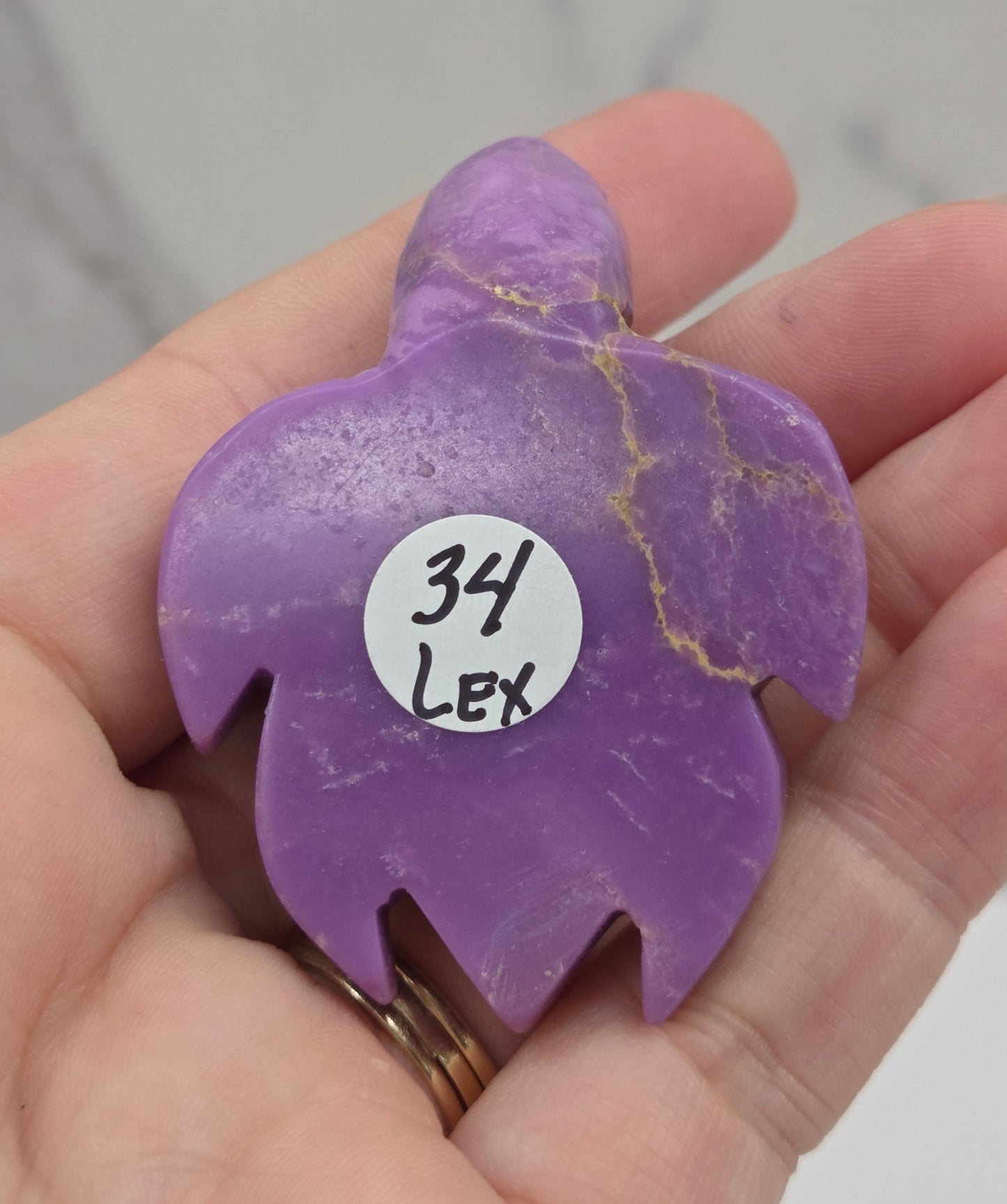 A hand holding a small purple carved stone charm with gold veining and a white circular sticker that reads '34 Lex'.