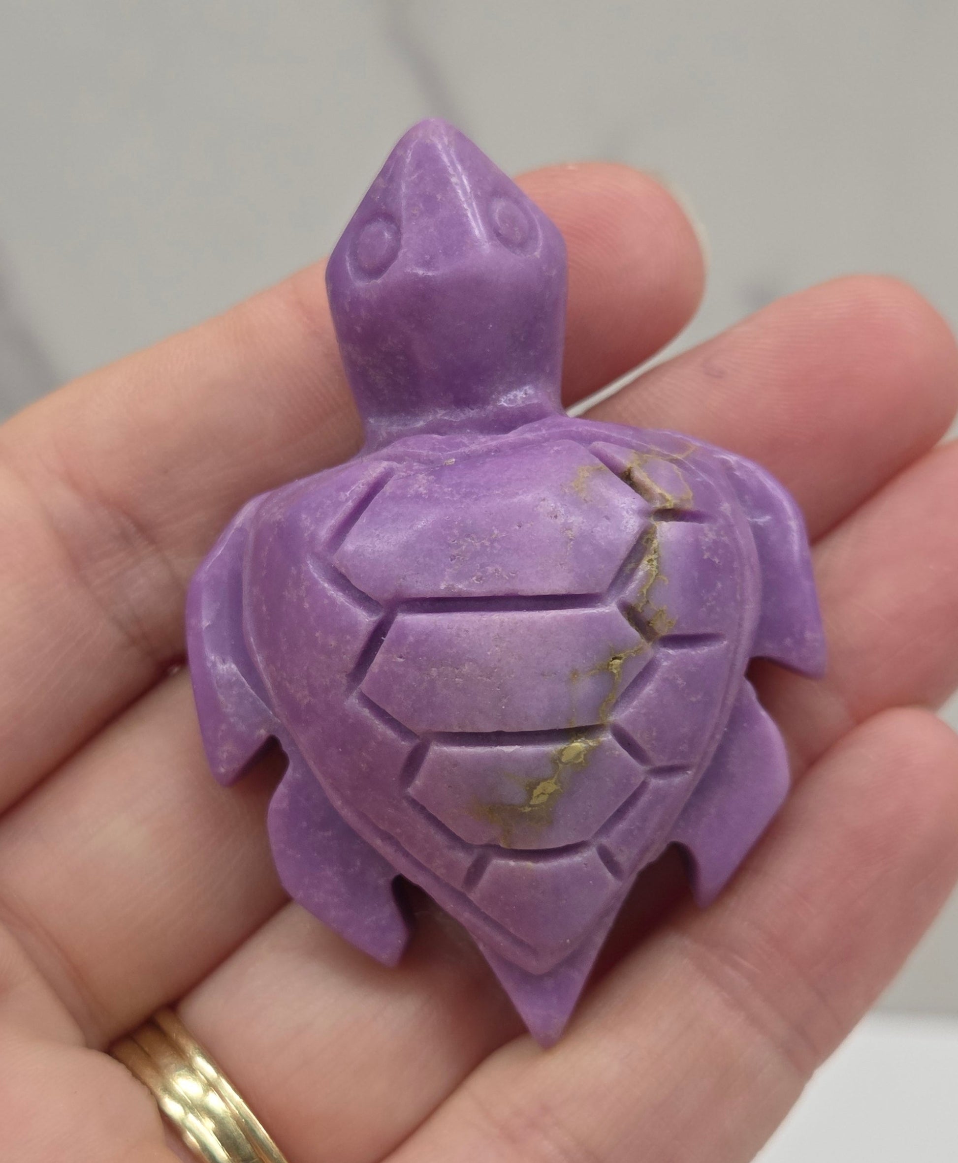 A small carved purple turtle figurine held in a person's hand, with a gold ring visible on the ring finger against a light marble-like background.