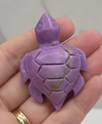 A small carved purple turtle figurine held in a person's hand, with a gold ring visible on the ring finger against a light marble-like background.