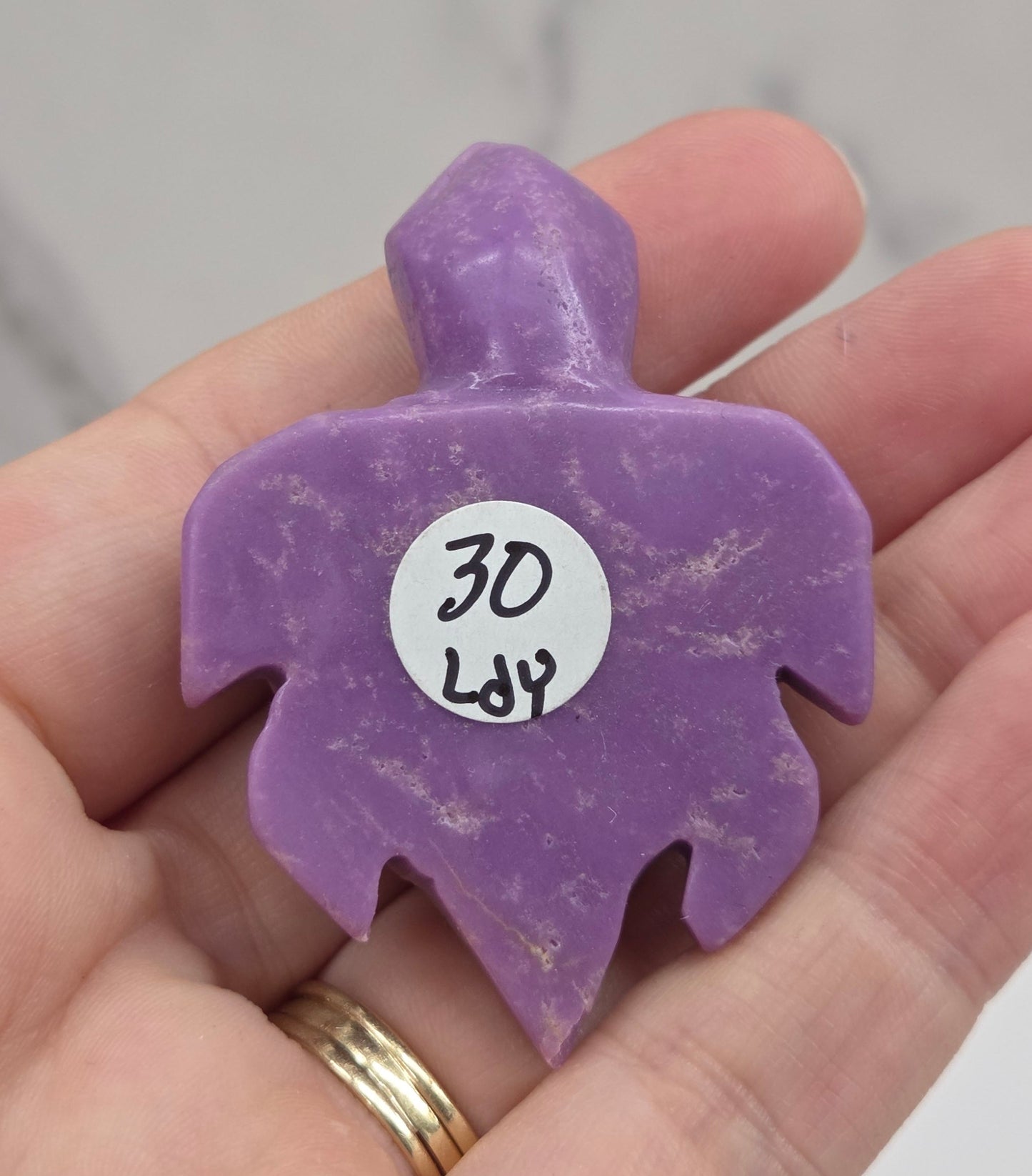 A hand holding a small purple carved stone or crystal with a white circular tag that reads '30 Lb'.