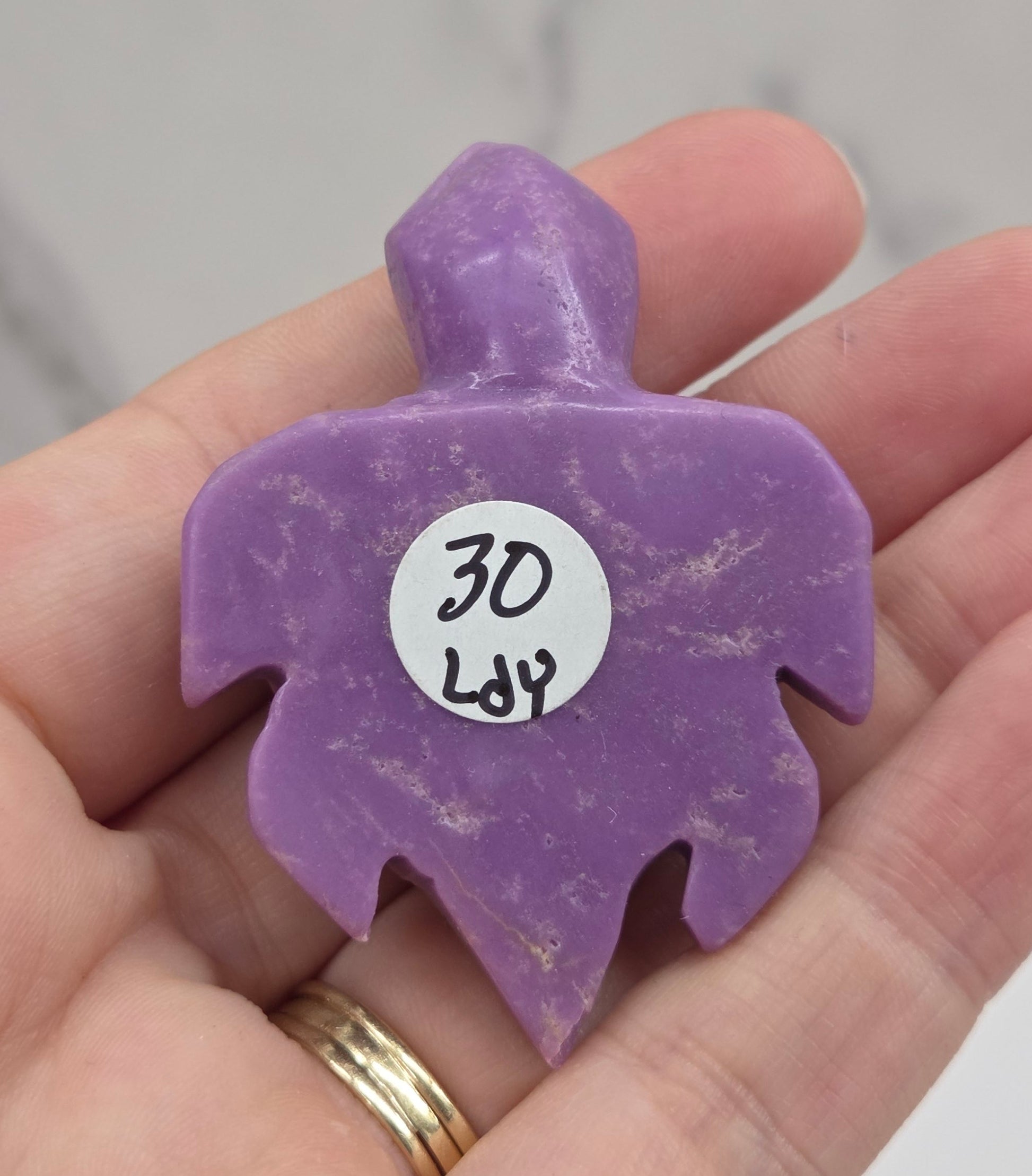 A hand holding a small purple carved stone or crystal with a white circular tag that reads '30 Lb'.