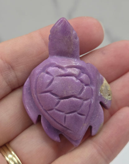 A small carved turtle-shaped pendant or charm in lavender purple, held in a person's hand. The turtle has a textured shell and a small gold accent on its back, and the hand is wearing a gold ring.