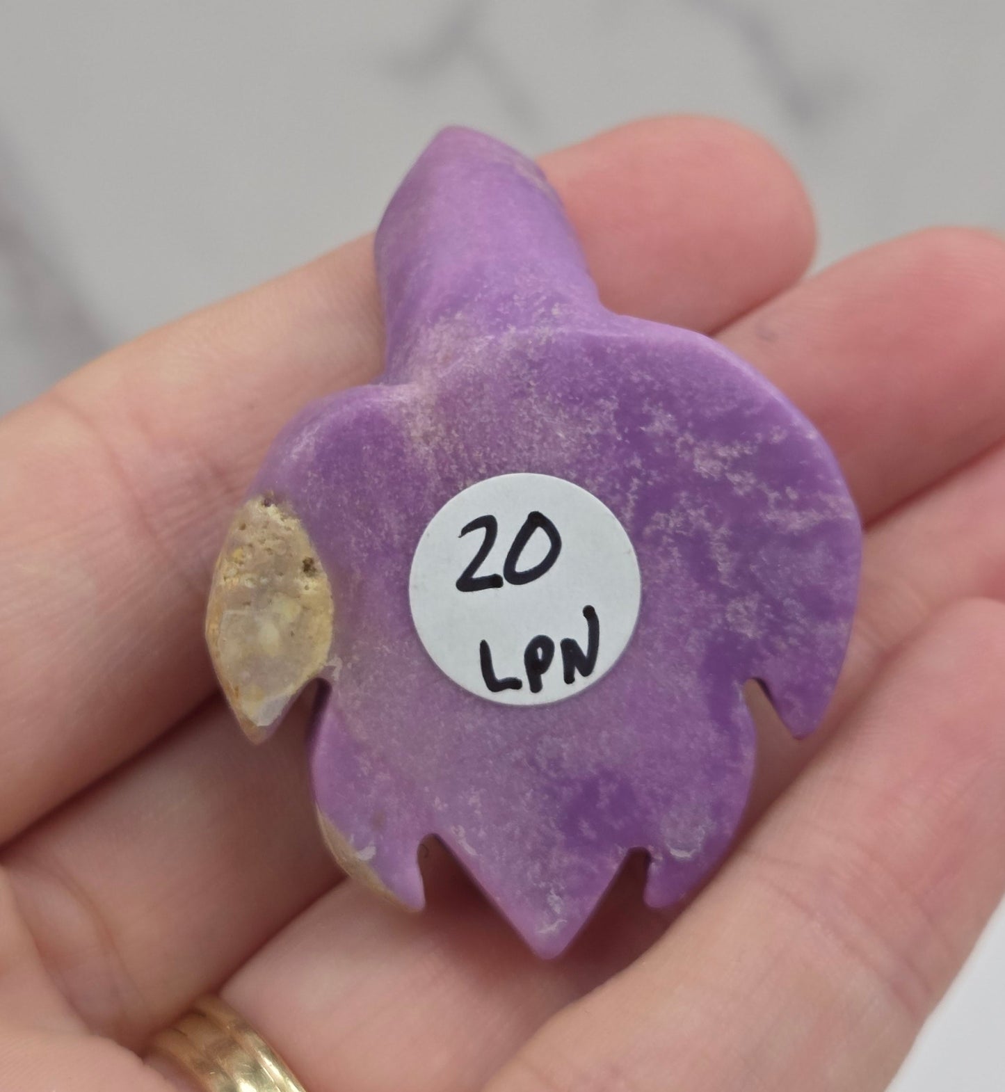 A hand holding a small purple decorative object with a white circular sticker that reads '20 LPM'. The background is a light gray marble surface.